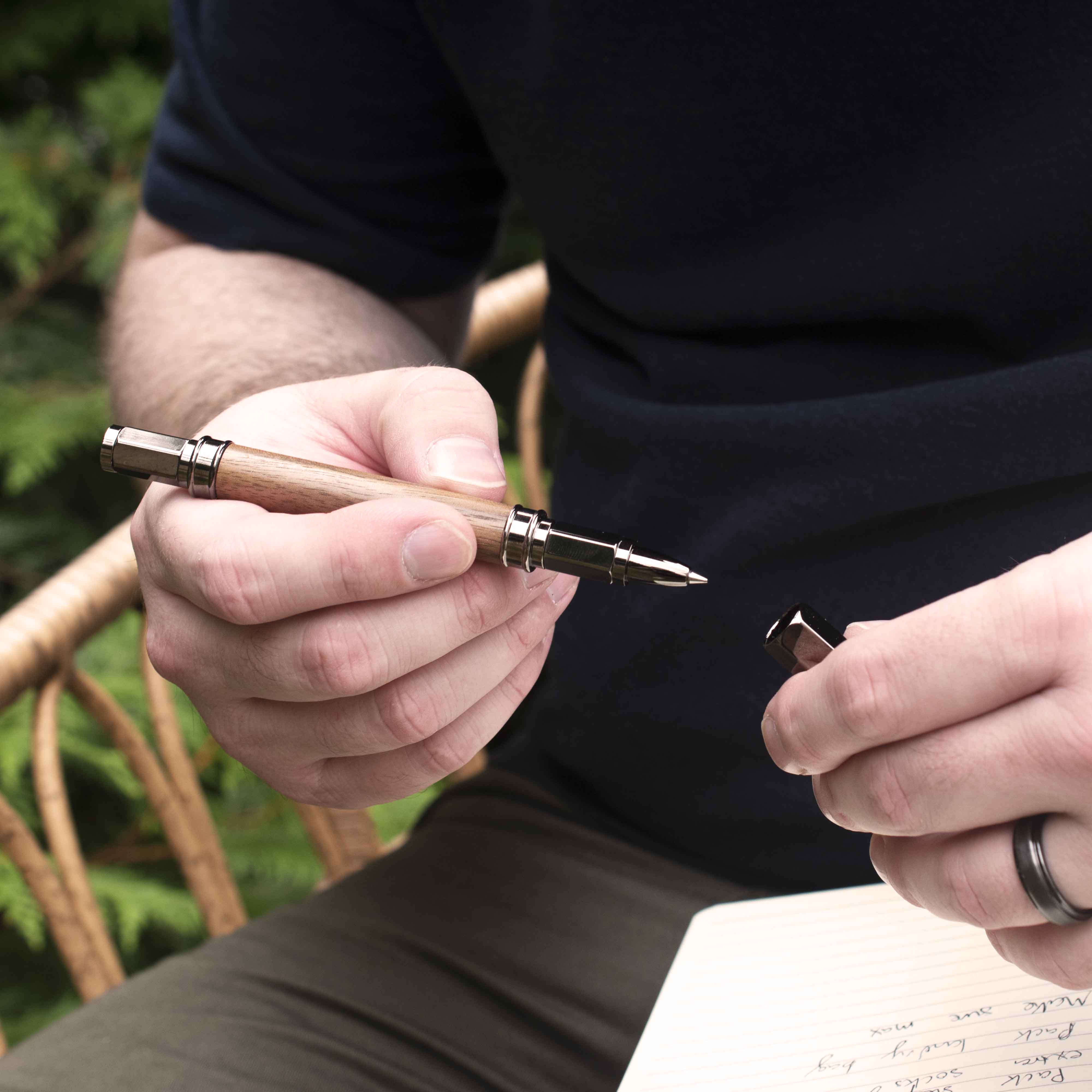 Black Walnut Rollerball Pen with the cap being put back on by someone seated in a whicker rocking chair outside in front of a fern tree