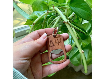 Hand holding a wooden keychain with a bottle opener and symbols against a green leafy background