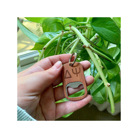 Hand holding a wooden keychain with a bottle opener and symbols against a green leafy background