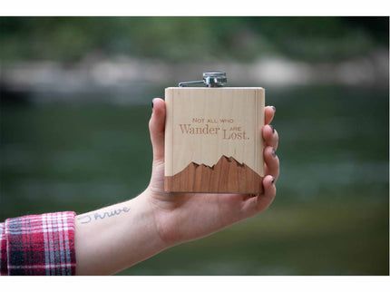 Hand holding a wooden flask with 'Not all who wander are lost' quote against a blurred natural background