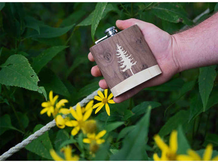 Hand holding a wooden flask with a tree design against a natural background with green leaves and yellow flowers.