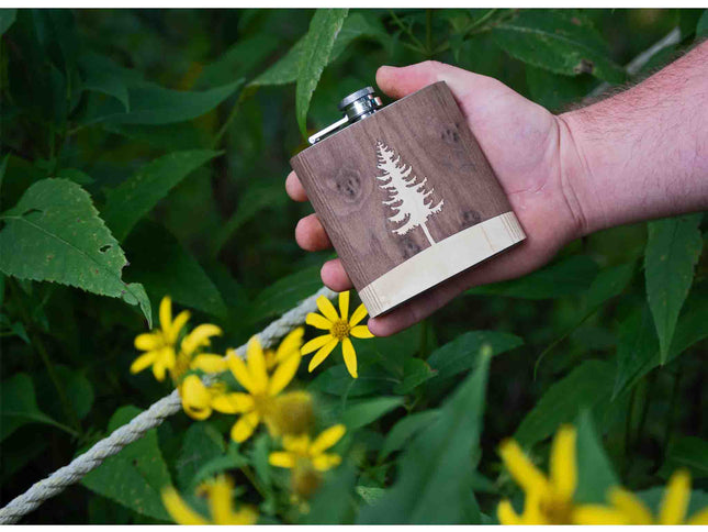 Hand holding a wooden flask with a tree design against a natural background with green leaves and yellow flowers.
