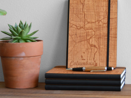Wooden notebook with map design and pen on a wooden surface next to a potted plant.