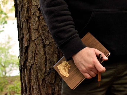 Person holding a wooden notebook and pen against a tree