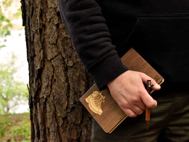 Person holding a wooden notebook and pen against a tree