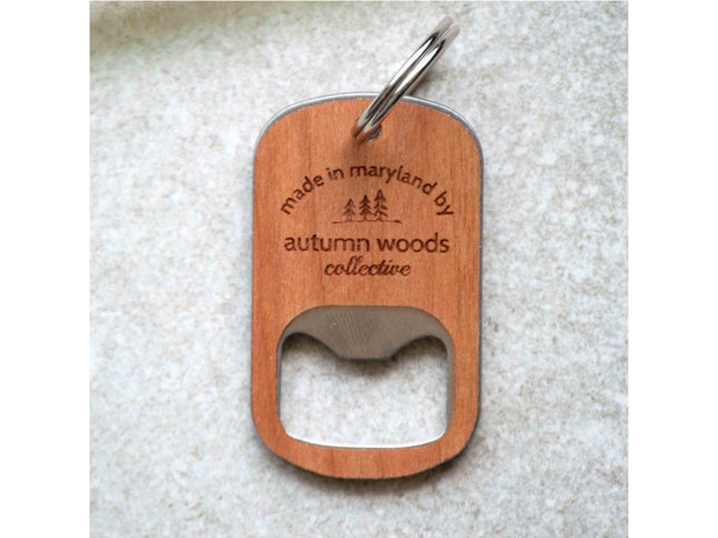 Wooden bottle opener keychain with 'made in maryland by autumn woods collective' text on a textured surface.