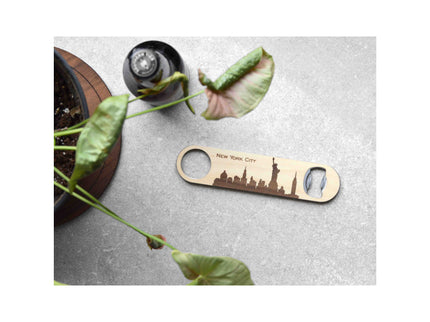 Bottle opener with New York City skyline design on a concrete surface with a plant.