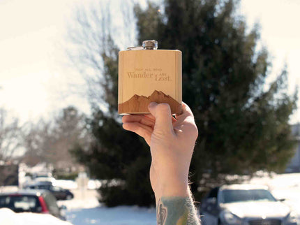 Person holding a wooden flask with 'Wander Lost' engraving outdoors on a snowy day.