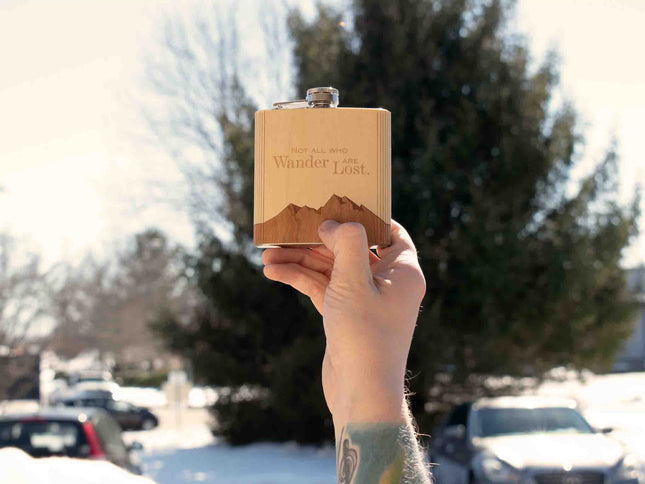 Person holding a wooden flask with 'Wander Lost' engraving outdoors on a snowy day.