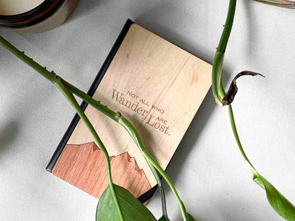Notebook with 'Not all who wander are lost' quote, surrounded by green leaves on a light background
