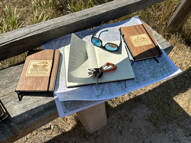 Two wooden journals, a notebook, sunglasses, keys, and a map on a wooden bench outdoors.