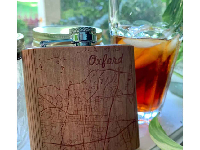 Wooden flask with 'Oxford' engraving on a table with a glass of amber liquid.