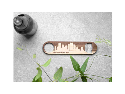 Wooden bottle opener with Pittsburgh, PA skyline design on a gray surface with a plant.