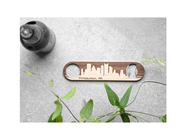 Wooden bottle opener with Pittsburgh, PA skyline design on a gray surface with a plant.