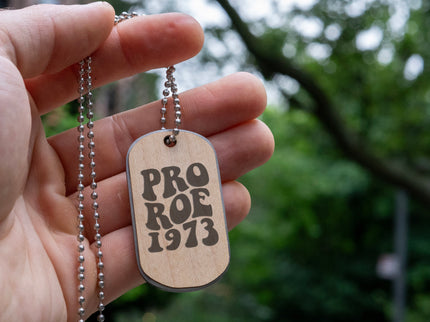 A hand holding a double sided maple wood dog tag with a stainless steel core and steel necklace chain engraved with "Pro Roe 1973" against a green tree background.