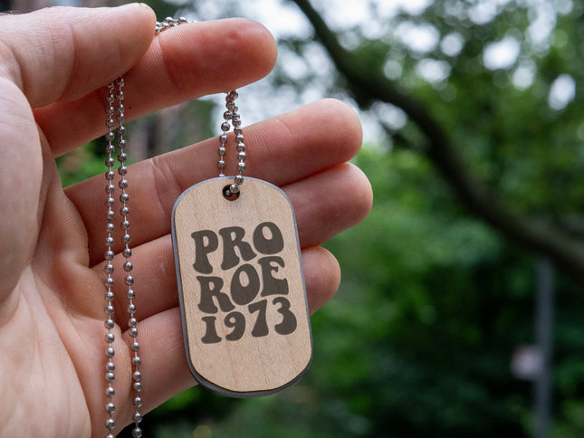 A hand holding a double sided maple wood dog tag with a stainless steel core and steel necklace chain engraved with "Pro Roe 1973" against a green tree background.