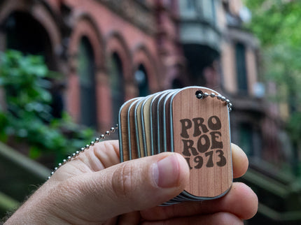 A hand holding a double-sided dog tag necklace with a stainless steel core and bead chain. The tag has a wood-like texture on one side and the text 'PRO ROE 1973' engraved on the other.