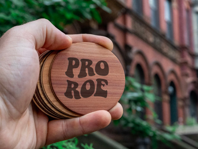 A hand holding a stack of round wooden stickers engraved with 'Pro Roe' against a street backdrop.