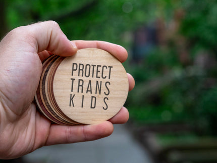A hand holding a protect trans kid round wooden sticker against a green street backdrop