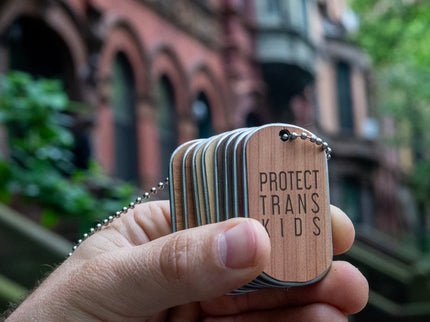 A hand holding a stack of double sided wooden dog tags with stainless steel cores and necklace chains, engraved with "Protect Trans Kids" against a street backdrop.