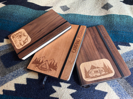 Three wooden notebooks with laser-engraved camping designs on a patterned fabric background
