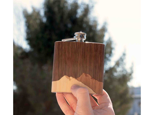 Hand holding a wooden flask with a mountain design against a blurred natural background