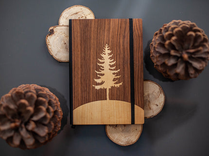 Wooden notebook with tree design surrounded by pine cones on a dark surface
