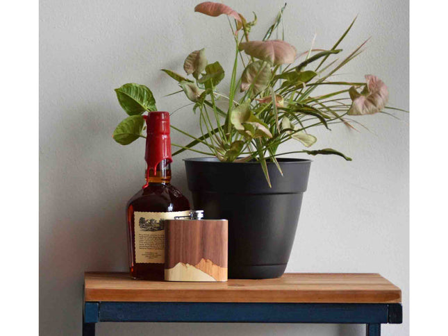 Bottle of whiskey, wooden flask, and potted plant on a wooden table against a plain wall.