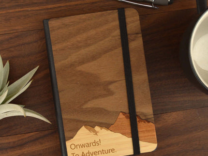 Wooden notebook with mountain design and 'Onwards! To Adventure.' text on a wooden surface with a pen and plant.