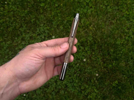 A handcrafted Equinox Rollerball Pen held in a person's hand, displaying a wooden body with a metal cap, placed against a grassy background.