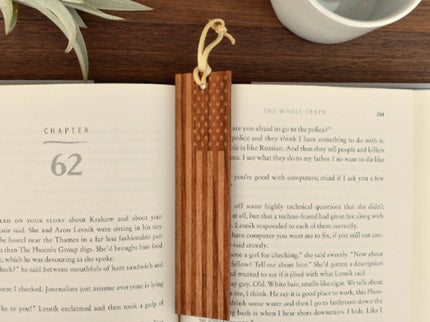 A real American Cherry wood bookmark with a leather loop, placed on an open book, showing the engraved American flag design.