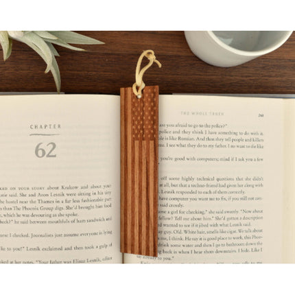 A real American Cherry wood bookmark with a leather loop, placed on an open book, showing the engraved American flag design.