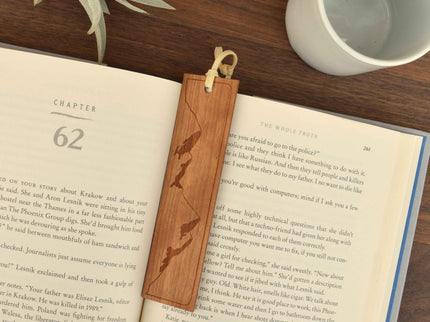 Wooden bookmark with a leather loop resting in a book. The bookmark is laser engraved with a mountain range.