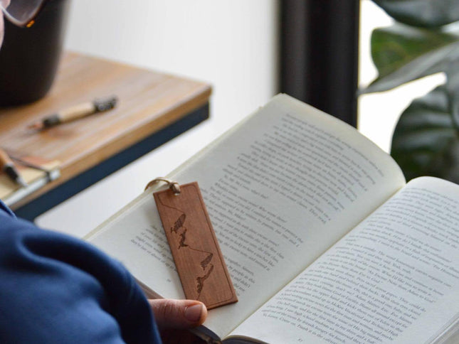 Person reading a book with a real american cherry wood bookmark engraved with a mountain range design.