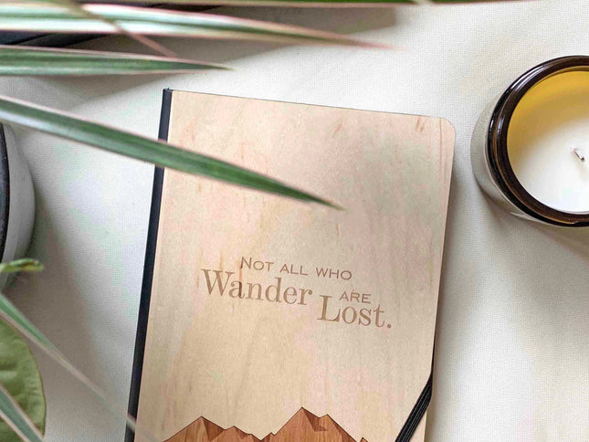A hardbound notebook with a wooden cover featuring an engraved mountain range and the text 'Not All Who Wander Are Lost'. It is placed on a white surface with a plant to the side and a candle in the background.