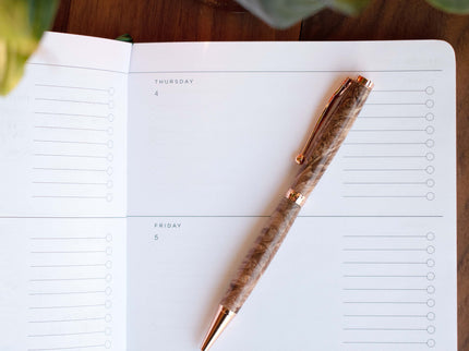 Marbled wood ballpoint pen with a golden colored pen kit laying on a journal page with a leafy background.